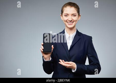 Waist up portrait of smiling businesswoman holding smartphone with blank screen to camera against grey background, copy space Stock Photo