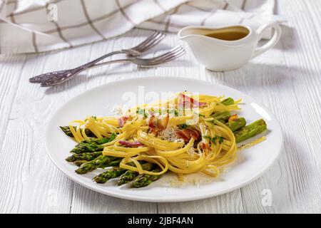 roasted asparagus spears with spaghetti sprinkled with shredded pecorino cheese, fried bacon pieces and parsley on white plate on white textured wood Stock Photo