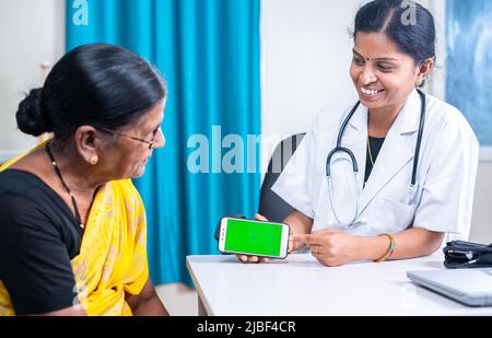 Doctor at hospital explaining or assisting senior woman patient by holding green screen mobile phone - concept of application, advertisement and Stock Photo