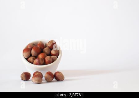 Hazelnuts, heap or stack of full nuts with shel in white cup and some on white background. Healthy and tasty vegan food, isolated and selective focus. Stock Photo