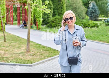 Stylish young blonde walks around the city and drinks a cold drink and talks on the phone. Time spent after work Stock Photo