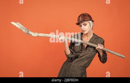 woman architect in protective helmet and boilersuit hold shovel on orange background, engineering Stock Photo