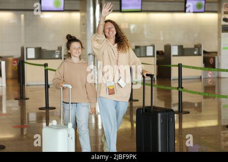 smiling young mother with teen daughter with suitcases is waving a hand to someone on meeting person after arrival plane an airport . Holiday vacation Stock Photo