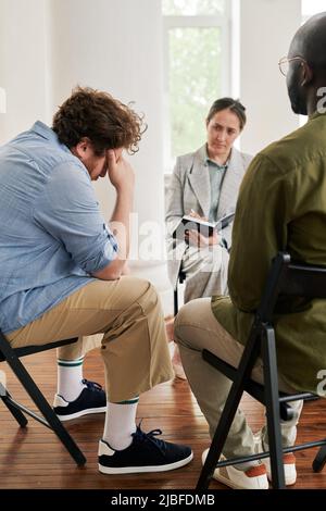 Fat young man touching his head while sitting in front of psychologist and describing his problem to her among other patients at session Stock Photo