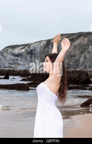 young brunette woman raising her arms on the beach in a white dress Stock Photo