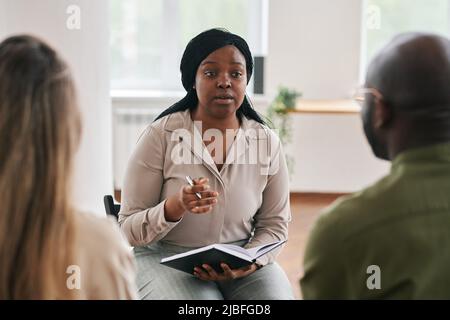 Young confident black woman with notebook and pen consulting intercultural couple sitting in front of her during session Stock Photo