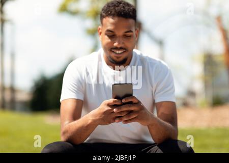 Mobile communication concept. Happy black guy using smartphone outdoors, sitting in parkland, selective focus Stock Photo