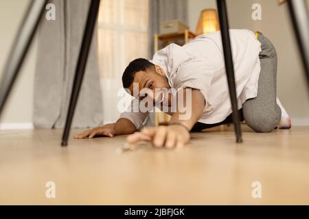 Young African American man trying to reach house keys, standing on his knees, dropping them on floor indoors Stock Photo