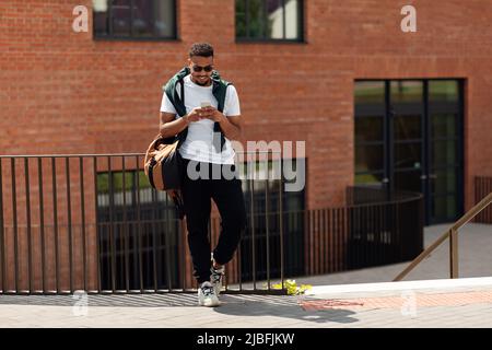 Trendy african american man surfing internet on cellphone, standing outdoor in city, texting on phone and smiling Stock Photo