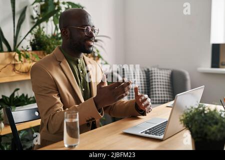 Young smiling black man in suit communicating woth patient during online psychological session while sitting in front of laptop Stock Photo