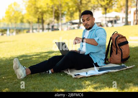 Young black man looking at smartwatch while working with laptop computer outdoors, sitting on blanket on grass Stock Photo