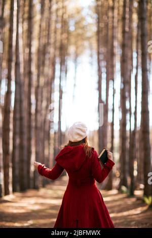 Back view of unrecognizable redhead female with outerwear and beret hat standing holding book in woods in fall Stock Photo