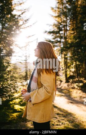 Side view of pensive young female tourist with long redheaded standing with eyes closed in casual clothes amidst autumn trees in Canada Stock Photo