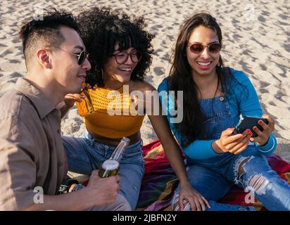 Happy multiracial man and women smiling and browsing social media on cellphones while resting on beach on sunny summer day on resort Stock Photo
