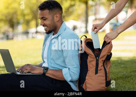 Female thief stealing smartphone from behind male bag, guy sitting on grass in park and working on laptop Stock Photo
