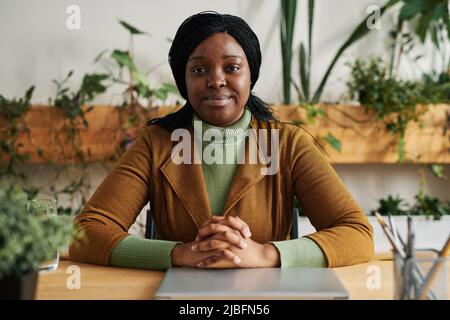 Young confident African American female psychologist in smart casualwear sitting by workplace in office and looking at camera Stock Photo