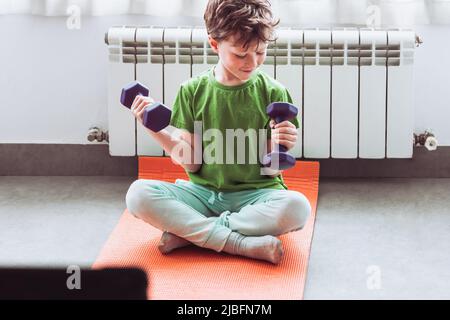 Merry child in activewear smiling and doing exercise with dumbbells while sitting cross legged on mat and watching tutorial video on tablet during onl Stock Photo