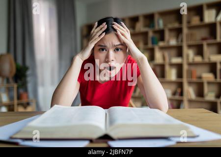 Sad shocked teen chinese lady student with open mouth study at table with book in room or library interior Stock Photo
