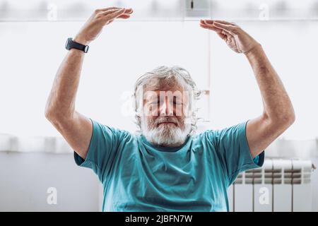 Elderly man in blue t shirt raising arms and meditating with closed eyes against window during yoga session in morning at home Stock Photo