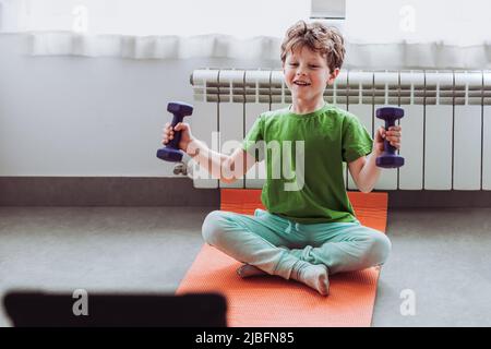 Merry child in activewear smiling and doing exercise with dumbbells while sitting cross legged on mat and watching tutorial video on tablet during onl Stock Photo