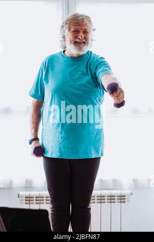 Happy senior bearded male in sportswear smiling and doing exercise with dumbbells during fitness workout in morning at home Stock Photo