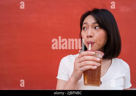Young Asian female with dark hair in casual clothes drinking iced coffee with straw from disposable cup and looking away against red wall on street Stock Photo