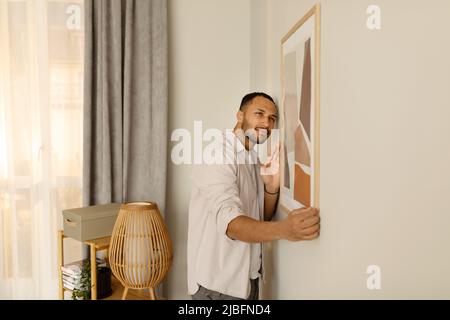 Portrait of handsome young African American man hanging picture frame on white wall at home, decorating his apartment Stock Photo