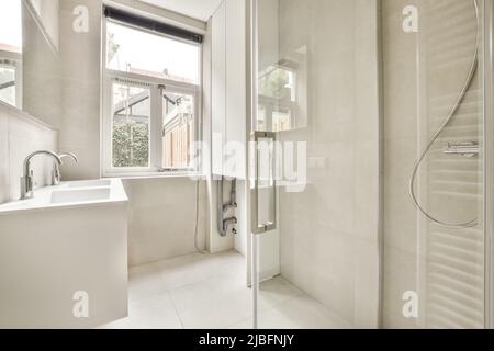 Cabinet with sink placed at wall in stylish bathroom with glass shower cabin and radiator on tilled walls with big windows Stock Photo
