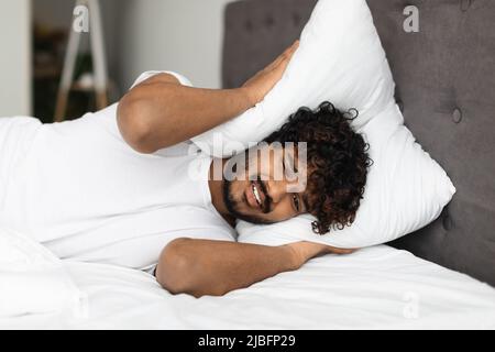 Angry eastern man laying in bed, covering head with pillow Stock Photo