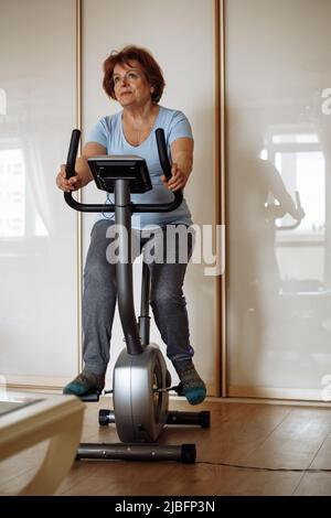 Portrait of elderly woman wearing blue T-shirt, grey trousers, sitting on stationary bicycle, working out exercises. Stock Photo
