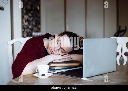 Portrait of tired middle-aged woman sitting at table near laptop, sleeping on crossed arms on pile of book sat home.  Stock Photo