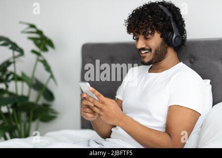 Happy indian guy sitting in bed with smartphone and headset Stock Photo