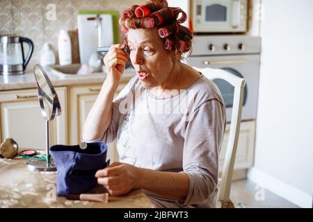 Elderly woman in pink hair-curlers, sitting at table, looking at mirror, putting powder rouge on skin with open mouth. Stock Photo