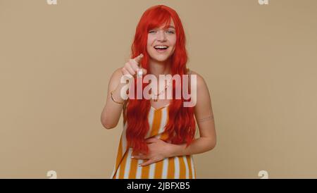 Amused ginger woman in tank top pointing finger to camera, laughing out loud, taunting making fun of ridiculous appearance, funny joke. Young red hair girl posing alone on beige studio wall background Stock Photo