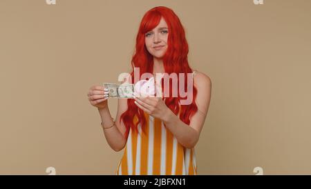 Poor woman upset over insufficient amount of money, holding piggybank and one dollar banknote. Financial crisis. Bankruptcy. Poverty and destitution. Young girl isolated on beige studio background