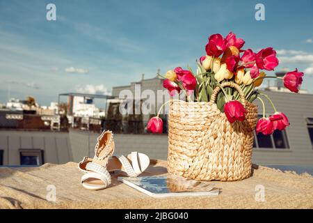 Composition of straw bag with bunch of pink, yellow flowers tulips, beige sandals and magazine on linen cloth on roof. Stock Photo