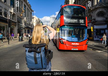 LONDON - May 18, 2022: Blonde female photographer taking photograph of red Double Decker Bus on busy London Street Stock Photo