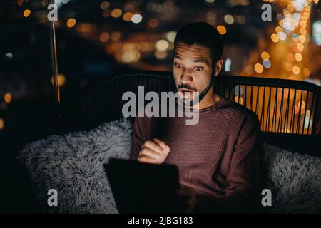 Young African AMerican man sitting on balcony with urban view and using tablet at night Stock Photo