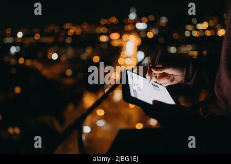 Close-up of man standing on balcony with urban view and using tablet at night Stock Photo
