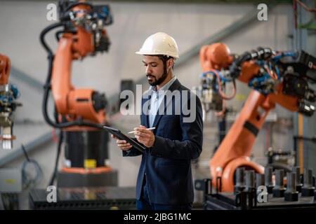Automation engineer uses tablet for programming robotic arm in factory. Stock Photo