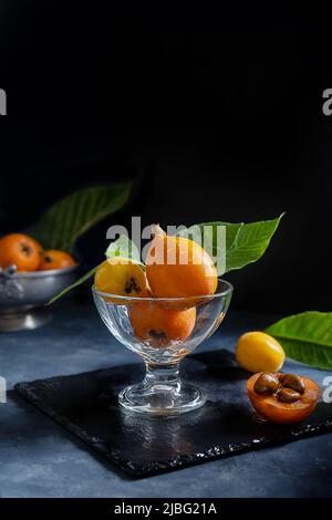 Yellow and orange Medlar fruits in glass cup over dark grey background. Chiaroscuro mood photo Stock Photo