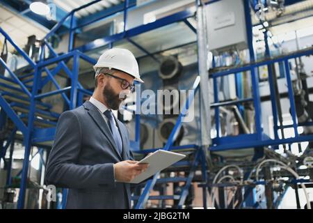 Chief Engineer in the hard hat walks through industrial factory while holding tablet. Stock Photo