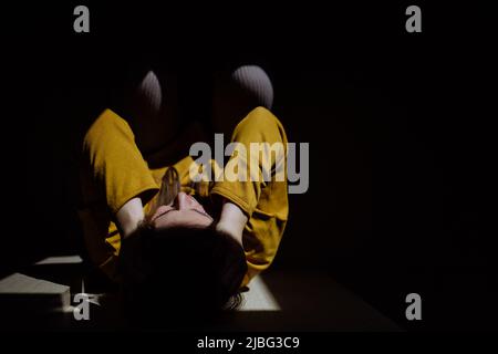 Woman suffering from depression and lying on floor with her hands covering head on black background. Stock Photo