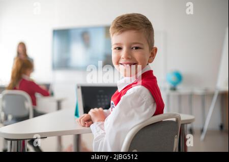 Happy schoolboy wearing school uniform sitting in class and smiling in the camera. Stock Photo