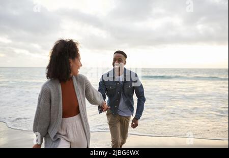 Smiling young multiethnic couple running hand in hand on a sandy beach Stock Photo