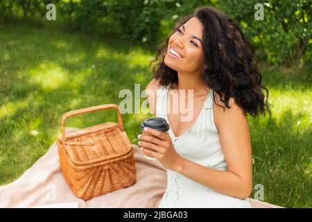 dreamy african american woman smiling and looking away while holding paper cup near picnic basket Stock Photo