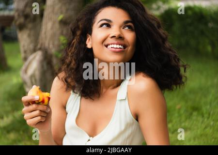 happy and young african american woman in white dress holding peach in park Stock Photo