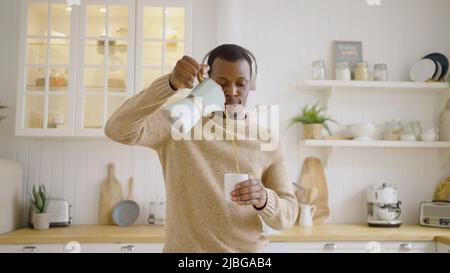 Cheerful man pours hot tea into cup from kettle dancing against countertop in kitchen. African American guy listens to dynamic music in headphones Stock Photo