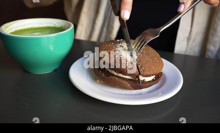Woman cuts brown cake with stainless steel cutlery in cafe. Plate with dessert and cup of green matcha served on table in cafe closeup Stock Photo