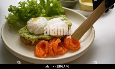 Chef puts trout slices on side of plate with chopsticks. Guacamole and poached eggs toast served on white plate with salad and fish slices closeup Stock Photo